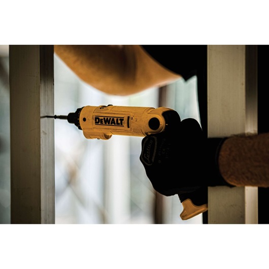 A close-up of a DEWALT electric screwdriver being used to drive a screw into a metal frame. The person is wearing gloves and working indoors.