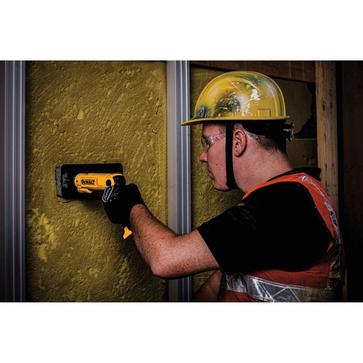 A worker wearing a yellow hard hat and safety vest uses a DEWALT DCF008 tool to work on a yellow surface wall at a construction site. The worker's face is blurred for privacy.