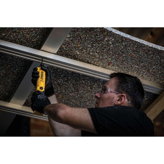 A person wearing gloves is using a yellow DEWALT DCF008 power tool to fasten a screw into a metal frame overhead. The person's face is blurred for privacy.