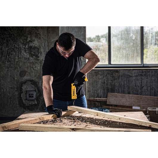 A person wearing black gloves uses a yellow DEWALT DCF008 cordless drill on a wooden frame in a workshop with concrete walls and a window.
