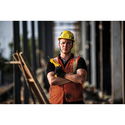 A construction worker wearing a yellow hard hat and orange safety vest stands with arms crossed, holding a DEWALT DCF008 tool in one hand at a building site. The worker's face is blurred for privacy.