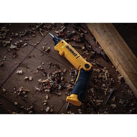 A yellow and black DEWALT cordless screwdriver tool lies on a wooden floor surrounded by wood shavings, screws, and a plank of wood.