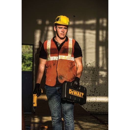 A person wearing a yellow safety helmet, an orange reflective vest, and gloves stands indoors holding a DEWALT-branded black toolbox in one hand and a yellow DEWALT power tool in the other hand. The background is a construction or industrial setting.