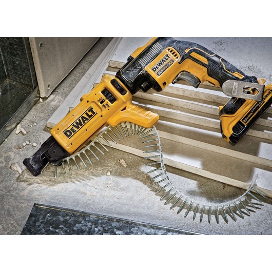 A DEWALT cordless drywall screw gun with an attached collated screw strip, lying on a construction site floor with drywall and debris nearby.