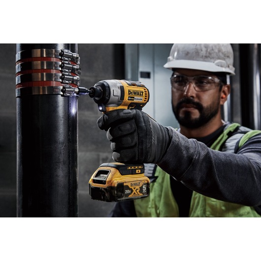 A person wearing work gloves and a safety vest is using a DEWALT 20V cordless impact driver to fasten metal clamps onto a large pipe. The person's face is blurred, and they are working indoors with industrial equipment.