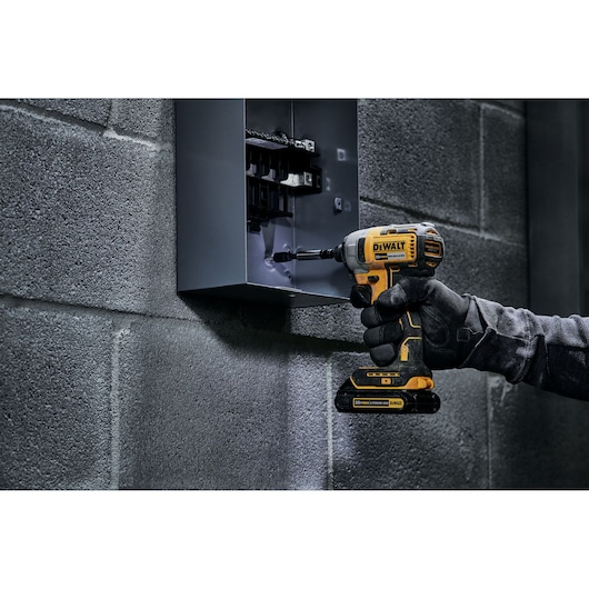 A person wearing black gloves uses a yellow and black DEWALT cordless impact driver (model DCF787C2) to work on an electrical panel mounted on a gray cinder block wall.