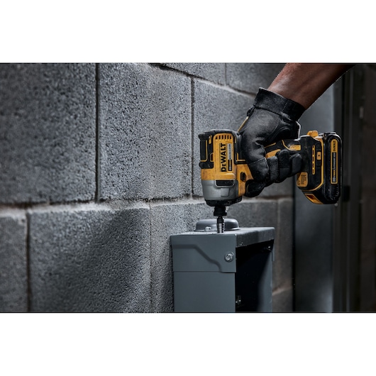 A person wearing black gloves uses a DEWALT cordless impact driver on a metal electrical box mounted on a gray cinder block wall.