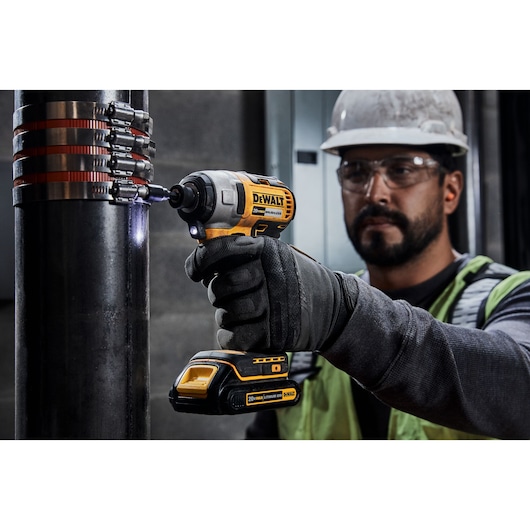 A worker wearing gloves and a safety helmet uses a DEWALT 20V cordless impact driver to tighten a clamp on a large metal pipe. The power tool model shown is DCF787C2.