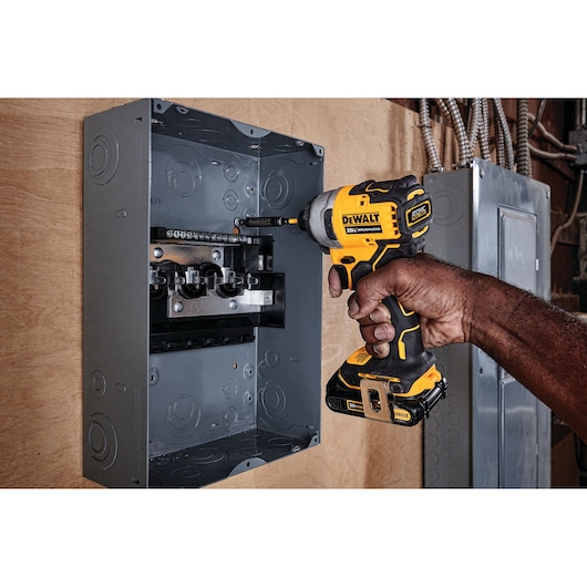 A person using a DEWALT cordless impact driver (model DCF809B) to work inside an electrical panel mounted on a wall.