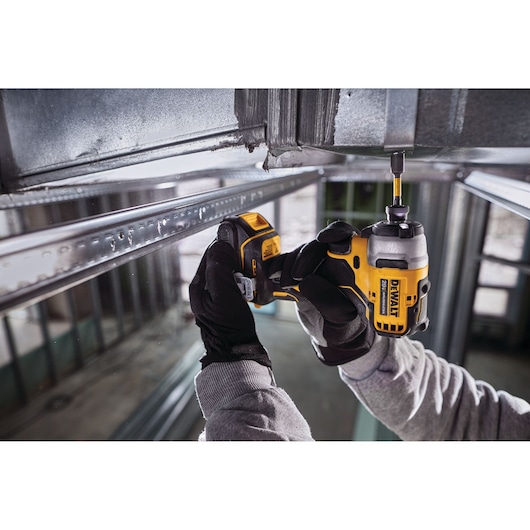 A person wearing gloves uses a DEWALT cordless impact driver to fasten a screw into a metal frame in an industrial setting.