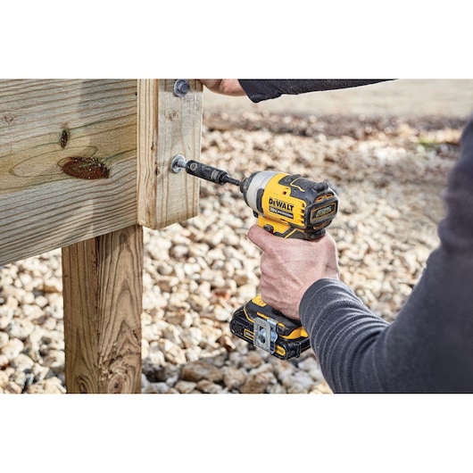 A person using a DEWALT cordless impact driver (model DCF809B) to drive a bolt into a wooden structure outdoors. The tool is yellow and black in color.