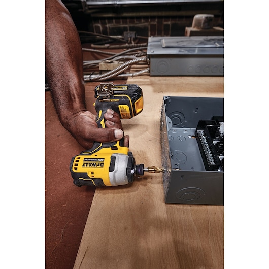 A person is using a DEWALT DCF809B 20V cordless impact driver to drill into a metal electrical box on a wooden surface.