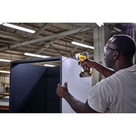 A person uses a DEWALT cordless power drill, model DCF809B, to assemble a large cabinet inside an industrial workshop with visible lighting and beams overhead.