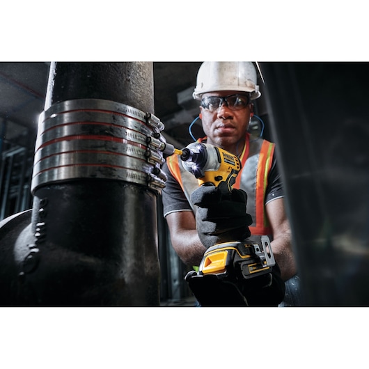 A person wearing safety gear and gloves is using a DEWALT DCF809B cordless impact driver to tighten industrial clamps on a large metal pipe in a construction or industrial setting.