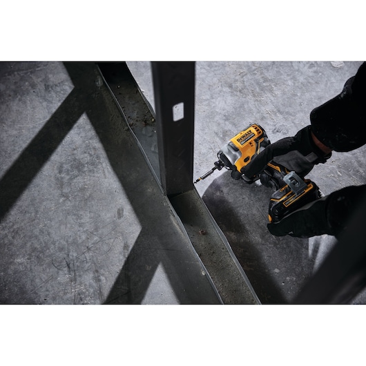A person wearing black gloves using a DEWALT cordless impact driver to drill a screw into a metal frame on a concrete floor.