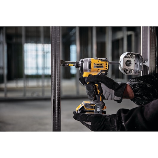 A person wearing black gloves is using a DEWALT 20V cordless impact driver (model DCF809B) to install a metal electrical box onto a steel framing stud at a construction site.