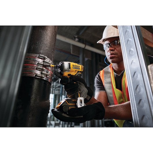A person wearing safety gear uses a DEWALT DCF809B cordless impact driver to fasten metal clamps onto a large industrial pipe at a construction site.