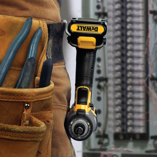 Close-up of a DEWALT cordless power tool hanging from a worker’s belt next to a tool pouch containing pliers, with an electrical panel in the background.