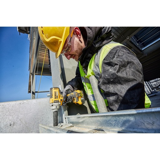 Worker uses DEWALT DCF894N impact wrench on metal at construction site, wearing helmet and safety vest.