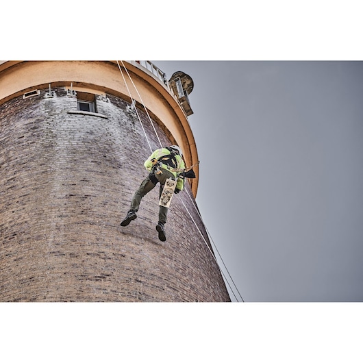 A worker wearing safety gear is hanging by ropes on the side of a tall brick building, using a DEWALT power tool.
