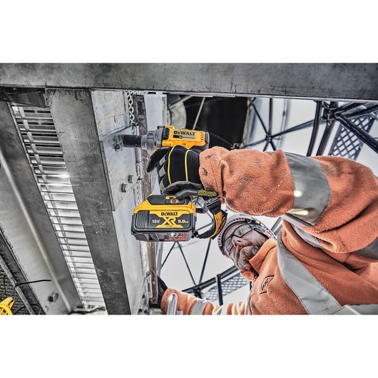 A worker wearing an orange safety jacket and gloves uses a DEWALT DCF894P2 cordless impact wrench to fasten bolts on a metal structure at an industrial worksite.
