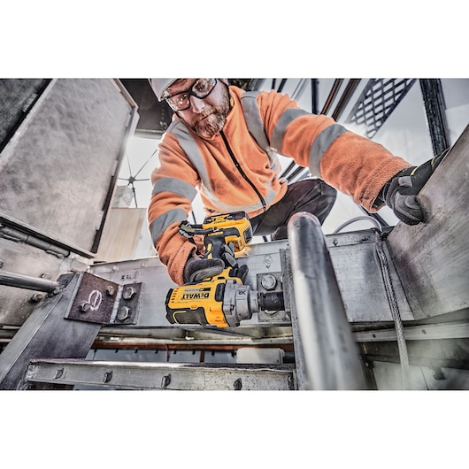 A person wearing an orange jacket and gloves is using a DEWALT DCF894P2 cordless impact wrench on a metal structure in an industrial setting.