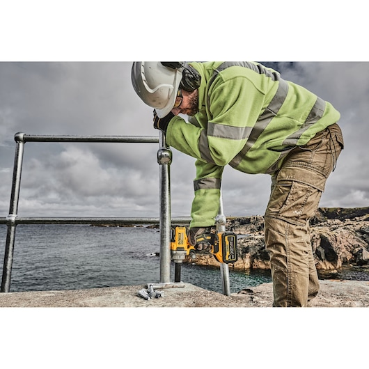 A person wearing safety gear, including a hard hat, high-visibility jacket, and safety glasses, uses a DEWALT cordless impact wrench (SKU: DCF894P2) to fasten a metal railing outdoors near rocky terrain and water.