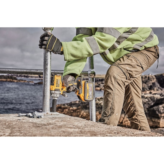 A person wearing work gloves and a high-visibility jacket is using a DEWALT cordless impact wrench (model DCF894P2) to secure a metal railing outdoors near a waterfront. Several bolts are lying on the concrete surface.