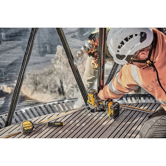 A person wearing a white safety helmet and orange workwear is using a DEWALT cordless impact wrench on a metal structure. The DEWALT DCF894P2 tool and battery are visible, along with a DEWALT measuring tape and screwdriver lying nearby.