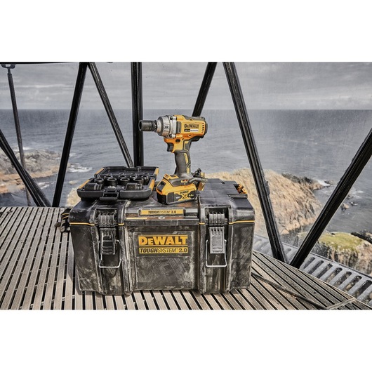 A DEWALT ToughSystem 2.0 toolbox with a DEWALT cordless impact wrench and accessories displayed on top, positioned in an industrial setting with an ocean view in the background.