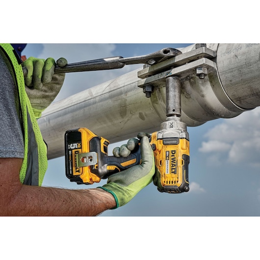 A person wearing safety gloves and a vest uses a DEWALT DCF894P2 cordless impact wrench to tighten or loosen a large bolt on a metal pipe outdoors. The tool is yellow and black, and is paired with a DEWALT 20V battery.
