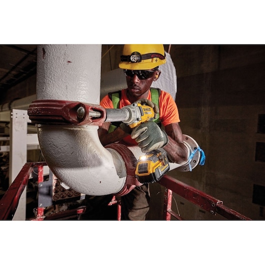 Worker uses DEWALT impact wrench on pipe while wearing safety gear at an industrial site.