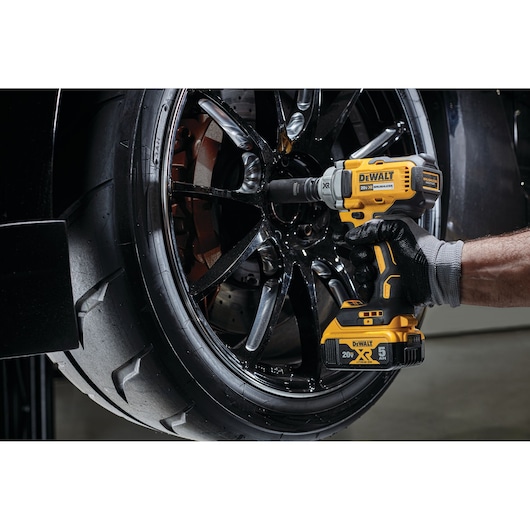 A gloved hand uses a yellow and black DEWALT cordless impact wrench to tighten or loosen bolts on a black car wheel.
