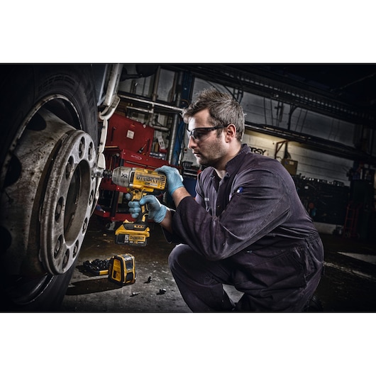 Person wearing a dark jumpsuit and blue gloves using a yellow DEWALT power tool to work on the wheel of a large vehicle. The scene is set in a workshop with various equipment visible in the background.
