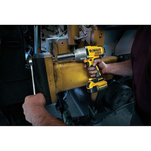 A person using a DEWALT DCF899P2 cordless impact wrench to tighten a large bolt on industrial machinery. The yellow and black power tool is held with one hand, while the other hand is using a wrench on the same bolt.