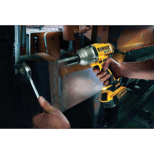 A person is using a DEWALT cordless impact wrench to fasten or loosen a large bolt, with another hand holding a wrench on the bolt in an industrial setting.