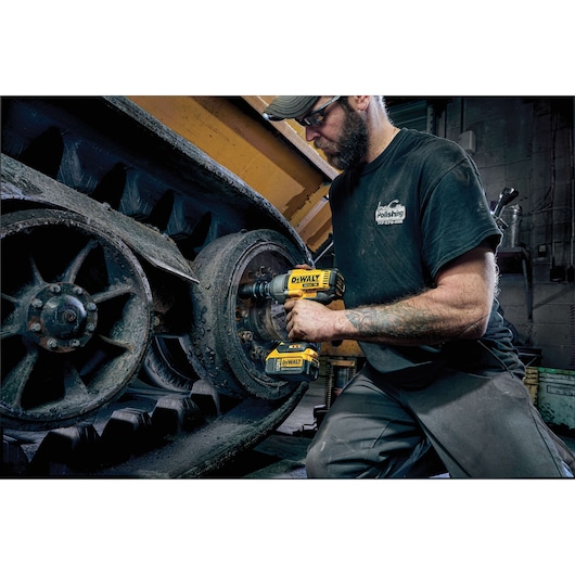 A person using a DEWALT DCF899P2 cordless impact wrench to work on the metal wheel of heavy industrial machinery. The person’s face is blurred for privacy, and the scene takes place in a workshop environment.