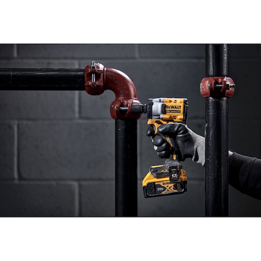A person wearing black gloves is using a DEWALT cordless impact wrench to tighten or loosen a red pipe fitting on black industrial pipes in front of a grey concrete wall.