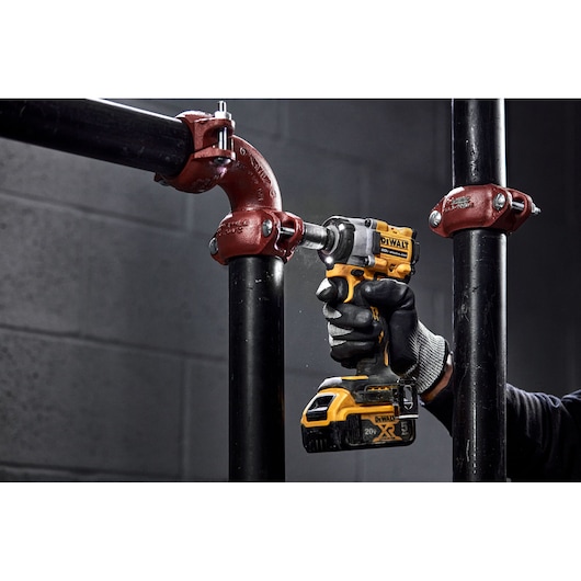 A person wearing gloves uses a DEWALT cordless impact wrench to tighten a bolt on a black metal pipe with red fittings in an industrial setting.
