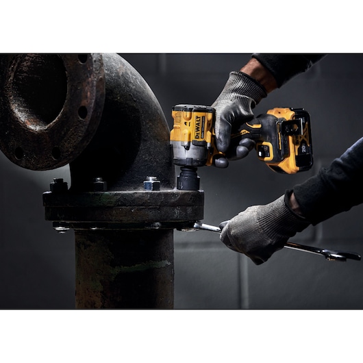A person wearing gloves uses a DEWALT cordless impact wrench to tighten bolts on a large industrial pipe flange. The scene is set in a workshop or industrial setting.
