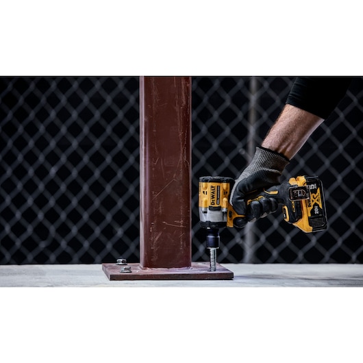 A person wearing a black glove uses a yellow DEWALT cordless impact wrench (model DCF922B) to tighten a bolt at the base of a brown metal post. The tool is positioned vertically against the bolt. The background features a metal fence with a grid pattern.