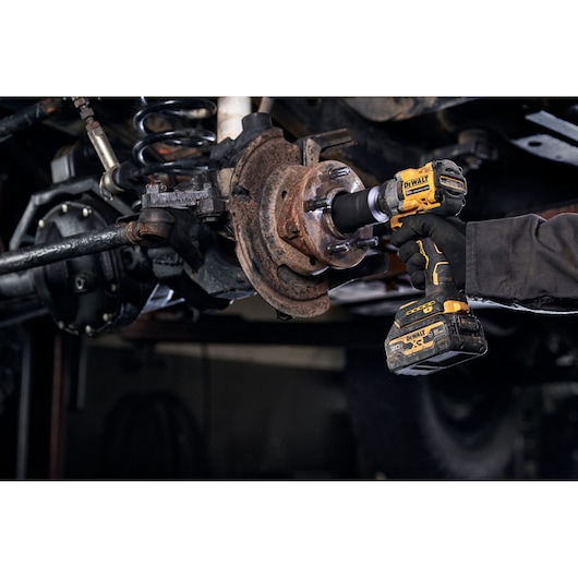 A person wearing gloves is using a DEWALT cordless impact wrench to work on a rusted automotive part under a vehicle.