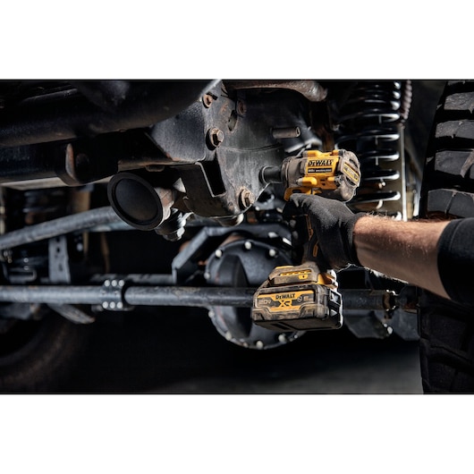 A person wearing gloves uses a yellow and black DEWALT cordless impact wrench (DCF922B model) to work on bolts under a vehicle. The tool is battery powered and designed for tough automotive tasks.