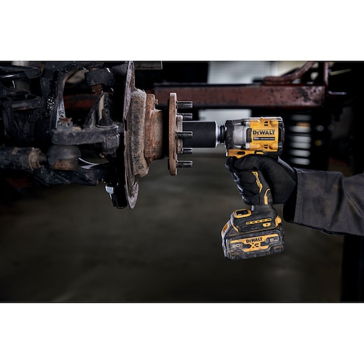 A person wearing gloves is using a DEWALT cordless impact wrench to work on a vehicle part, likely a wheel hub or brake assembly, in a workshop setting.