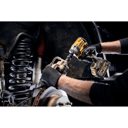 A person wearing black gloves uses a yellow DEWALT cordless impact wrench to work on a vehicle's brake assembly, with visible suspension components in the background.
