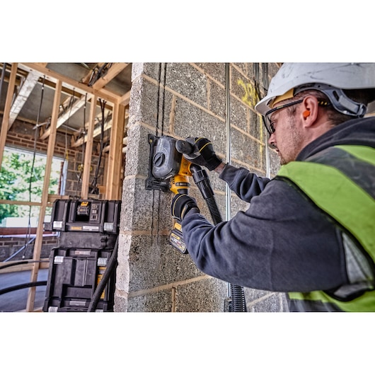 A person wearing safety equipment uses a DEWALT DCG200T2 tool to cut into a concrete wall at a construction site. Stacks of DEWALT storage boxes are seen in the background.