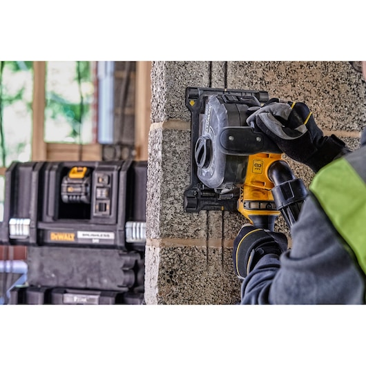 A person wearing work gloves uses a DEWALT wall chaser tool to cut grooves into a concrete block wall. Multiple DEWALT toolboxes are visible in the background.