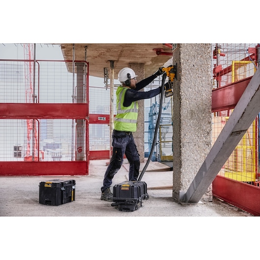A construction worker wearing safety gear, including a helmet and high-visibility vest, uses a DEWALT power tool to cut into a concrete pillar at a construction site. DEWALT storage and equipment boxes are positioned on the ground nearby.