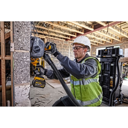 A worker wearing a reflective vest and hard hat operates a DEWALT DCG200T2 power tool to cut or grind a concrete wall in a construction site. The tool is connected to a vacuum hose to control dust. Construction materials and equipment are visible in the background.
