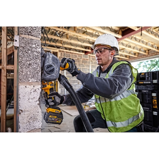 Person wearing a safety helmet and high-visibility vest using a DEWALT cordless wall chaser tool on a concrete block wall at a construction site. The tool is equipped with a dust extraction hose. DEWALT storage boxes are visible in the background.